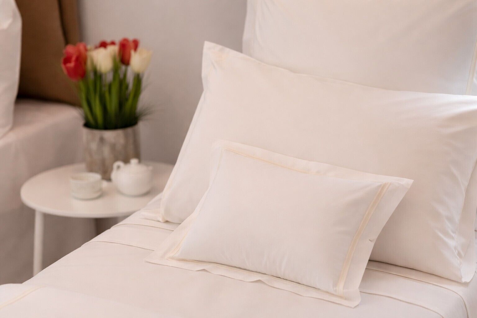 A bed with white bedding and pillows, a small round table with a vase of red and white tulips, and a white teapot.