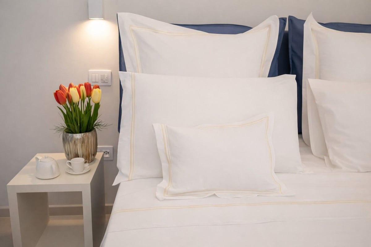 A neatly made bed with white bedding and pillows, a bedside table with a white teapot and cup, and a vase with red and yellow tulips.