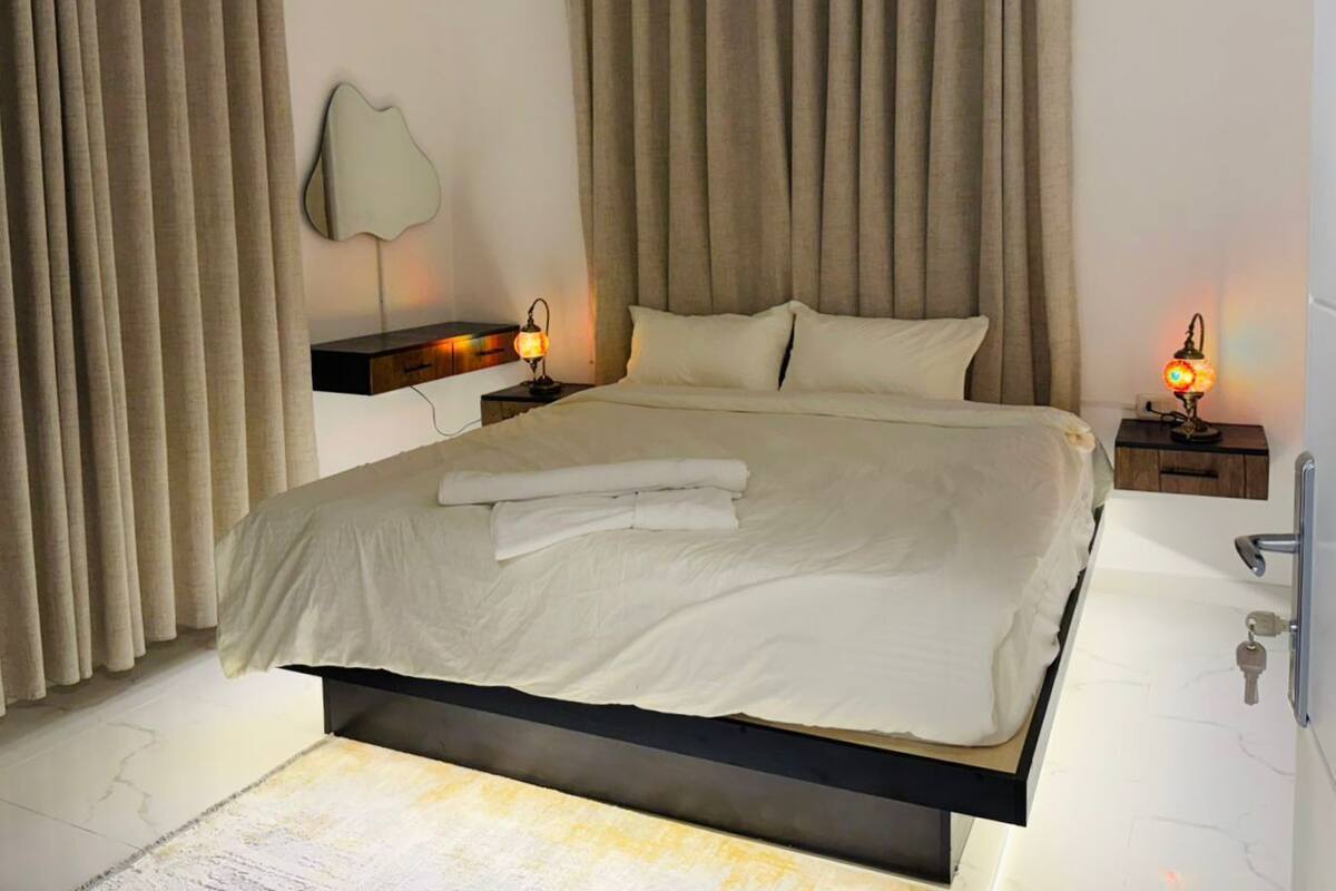 A modern bedroom with a large bed, two bedside tables with lamps, and a wall-mounted mirror.