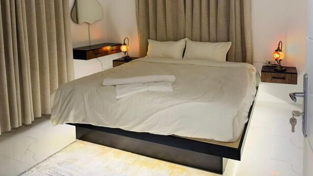 A modern bedroom with a large bed, beige curtains, and bedside lamps. - Beit Ahlan Suites (Amman)