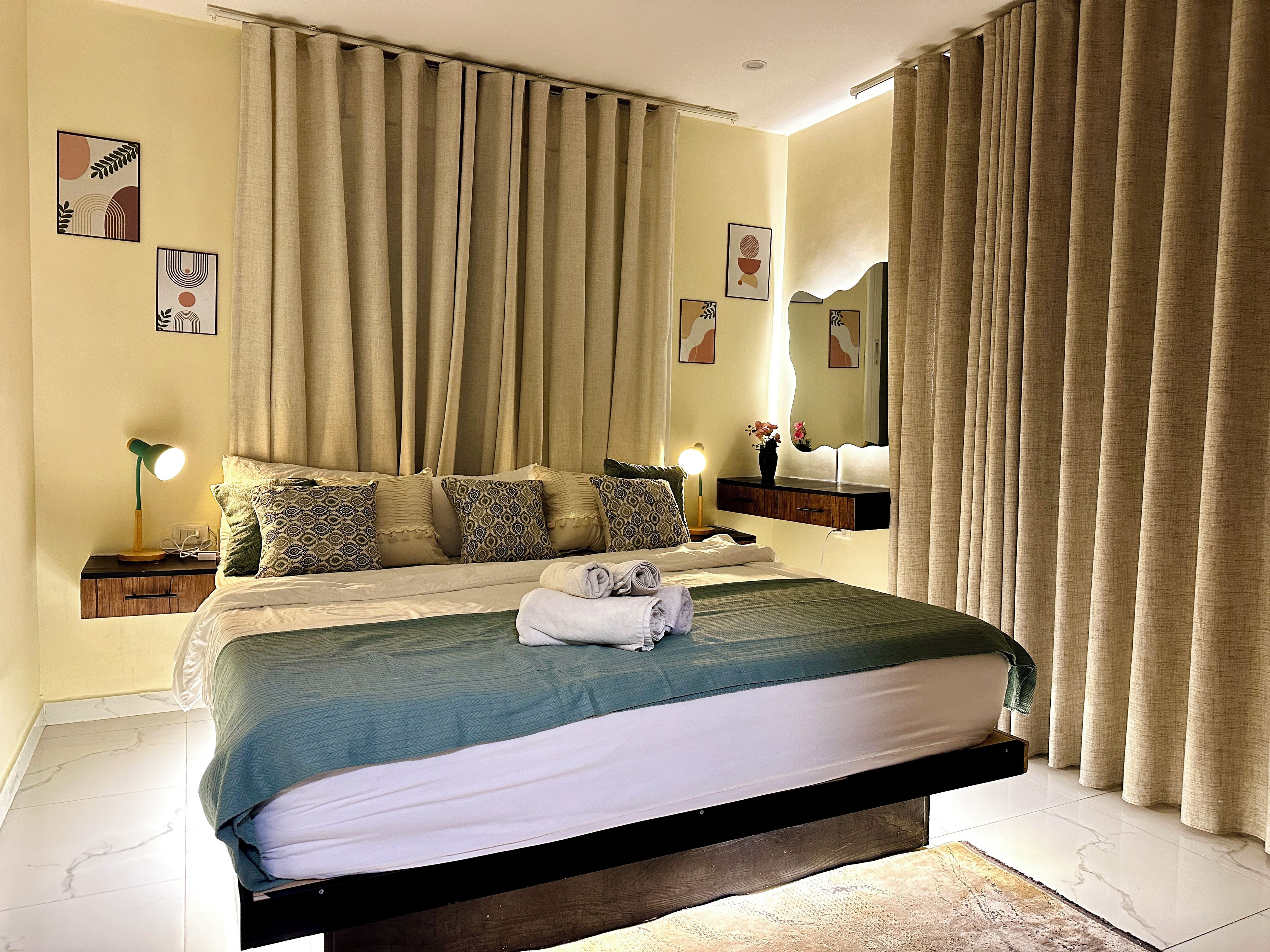 Cama bem arrumada com colcha verde e branca, luminária de cabeceira e mesa de cabeceira com abajur.