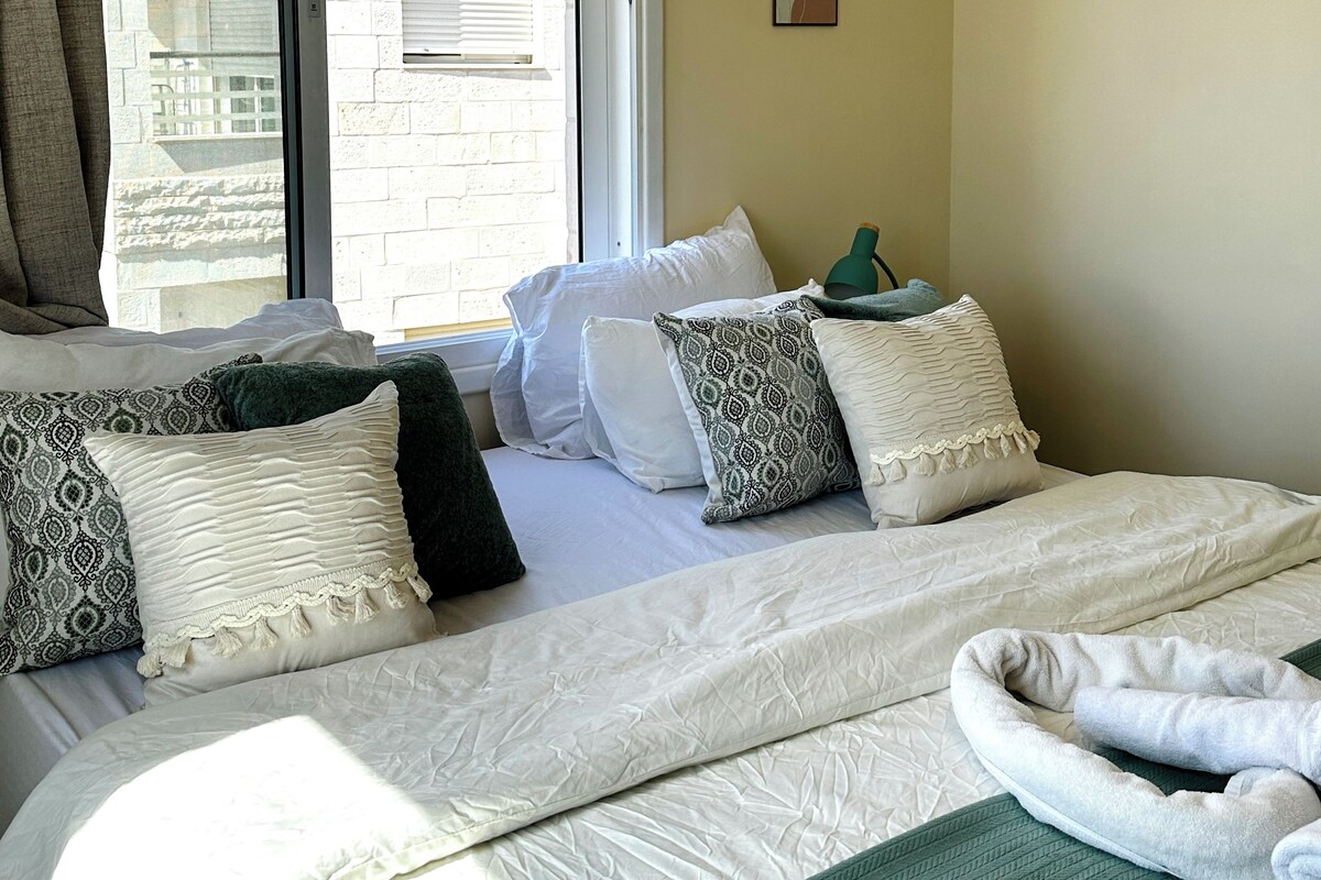 A bedroom with a bed, pillows, a green blanket, and a window with blinds.