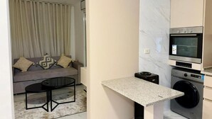 A compact living space with a sofa, a coffee table, a small kitchen area including a microwave and washing machine, and a marble backsplash. - Beit Ahlan Suites (Amman)