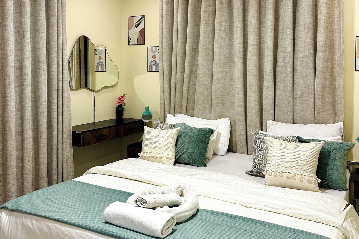 A double bed with a green and white bedspread, two pillows, and a rolled towel.