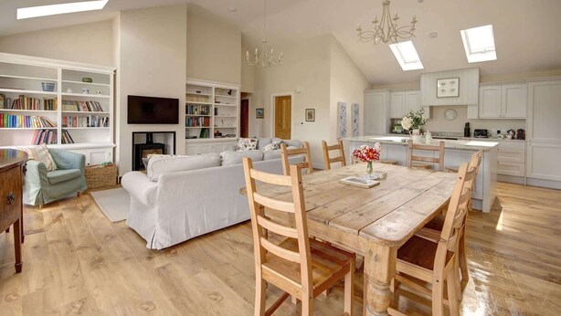 Een ruime woonkamer met een houten eettafel, witte banken en een open haard. Er zijn boekenkasten, een televisie en een keuken te zien.