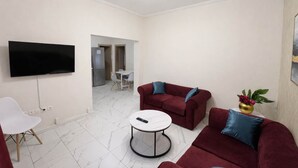 Een woonkamer met een flatscreen televisie, twee rode banken met blauwe kussens, een witte salontafel en een kleine bijzettafel met een vaas.