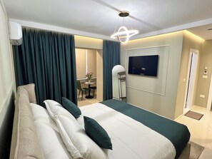 Una habitación de hotel moderna con una cama grande, un televisor montado en la pared y un área de comedor visible a través de las cortinas.