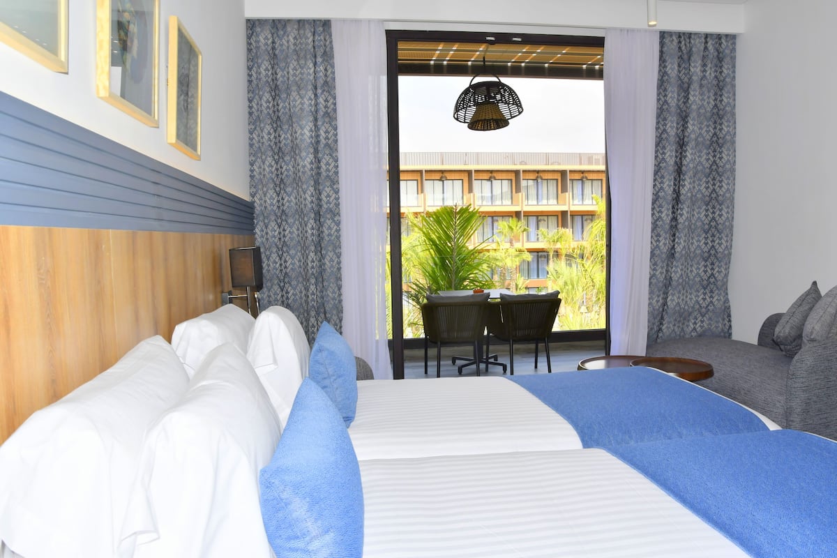 A hotel room with a bed, a sofa, a dining table with chairs, and a view of a building outside.