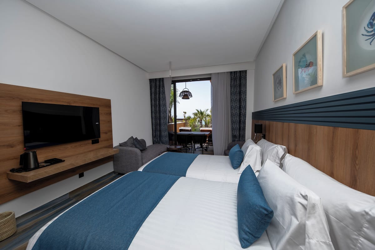 A hotel room with a TV, two beds, a sofa, a dining table, and a view of the outdoors.