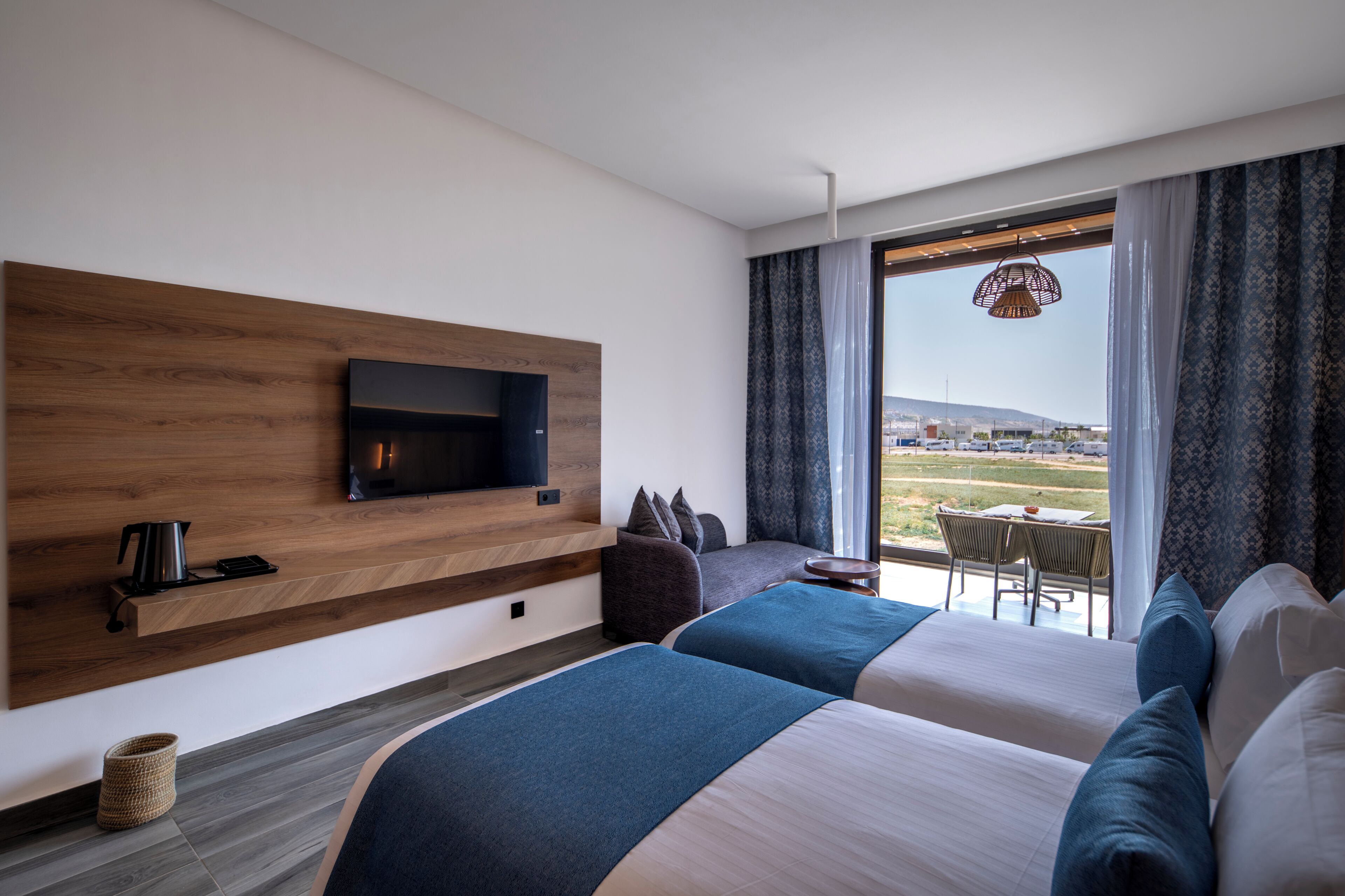 A modern hotel room with a large bed, a TV mounted on a wooden panel, a small table with chairs, and a view of the outdoors.