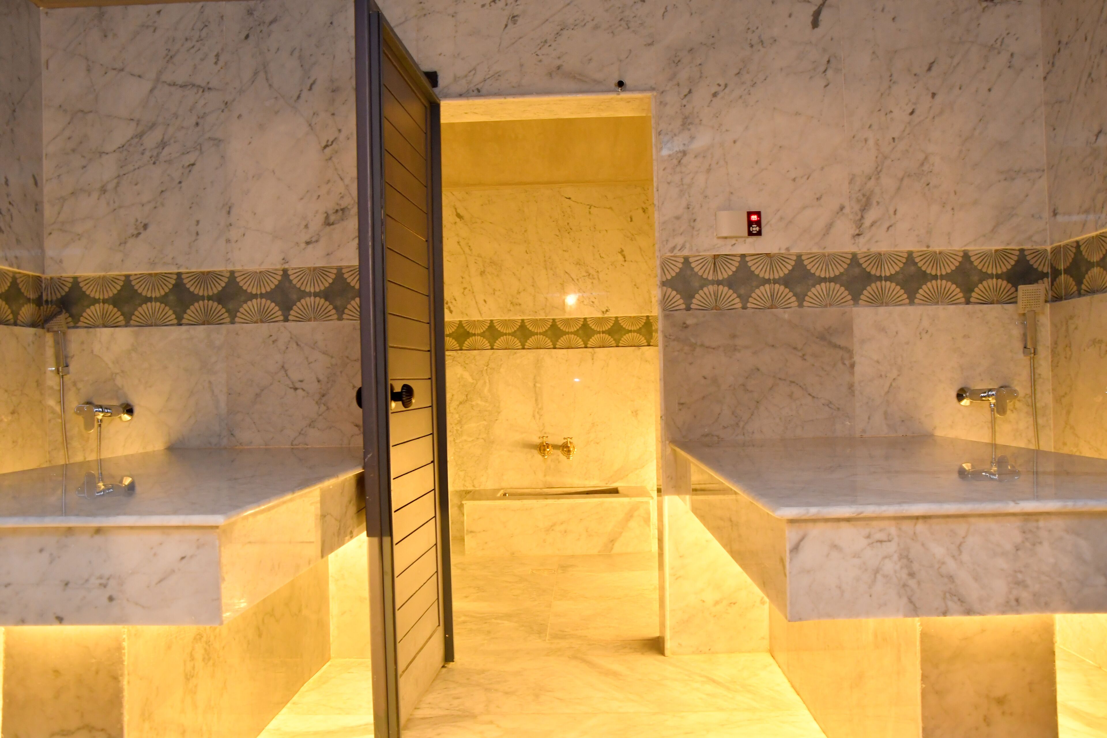 Un baño con paredes de mármol, una zona de ducha con azulejos amarillos y dos lavamanos con grifos.