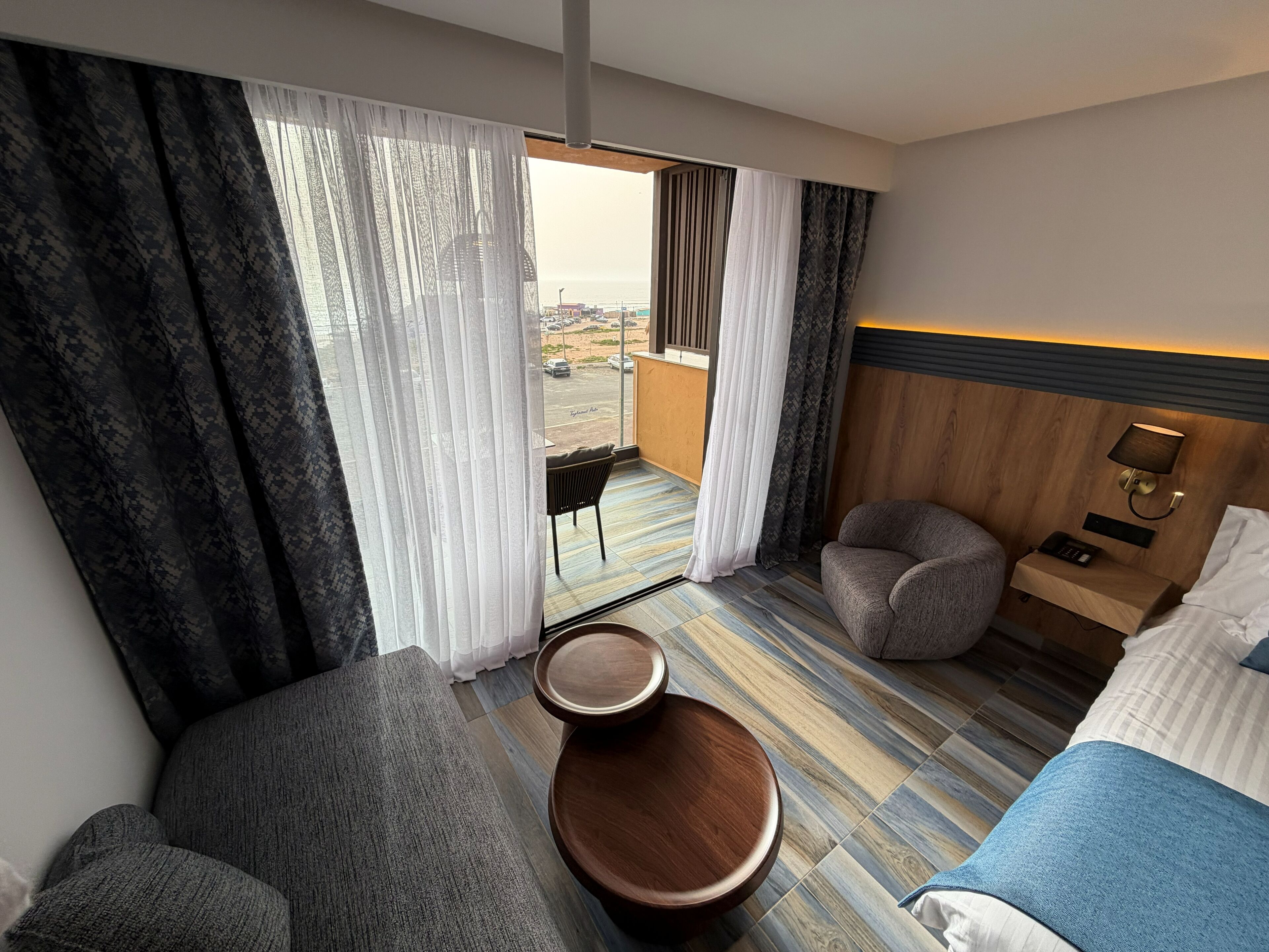 A hotel room with a bed, a seating area with a table, a view through a window, and a nightstand with a lamp.