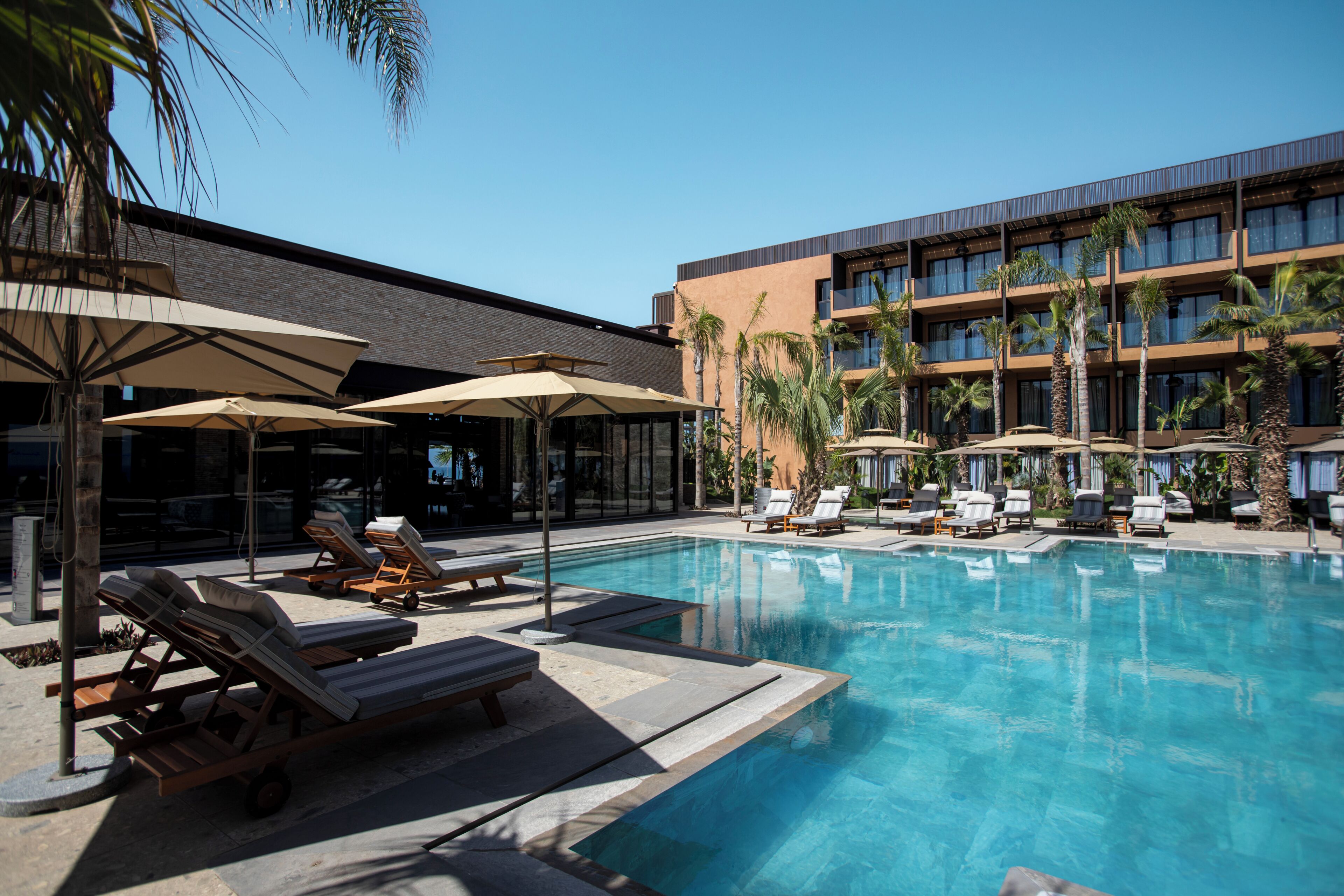 A pool area with lounge chairs and umbrellas, a building with multiple floors and balconies, and palm trees.