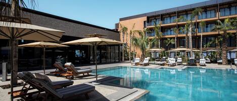 A pool area with lounge chairs and umbrellas, a building with multiple floors and balconies, and palm trees.