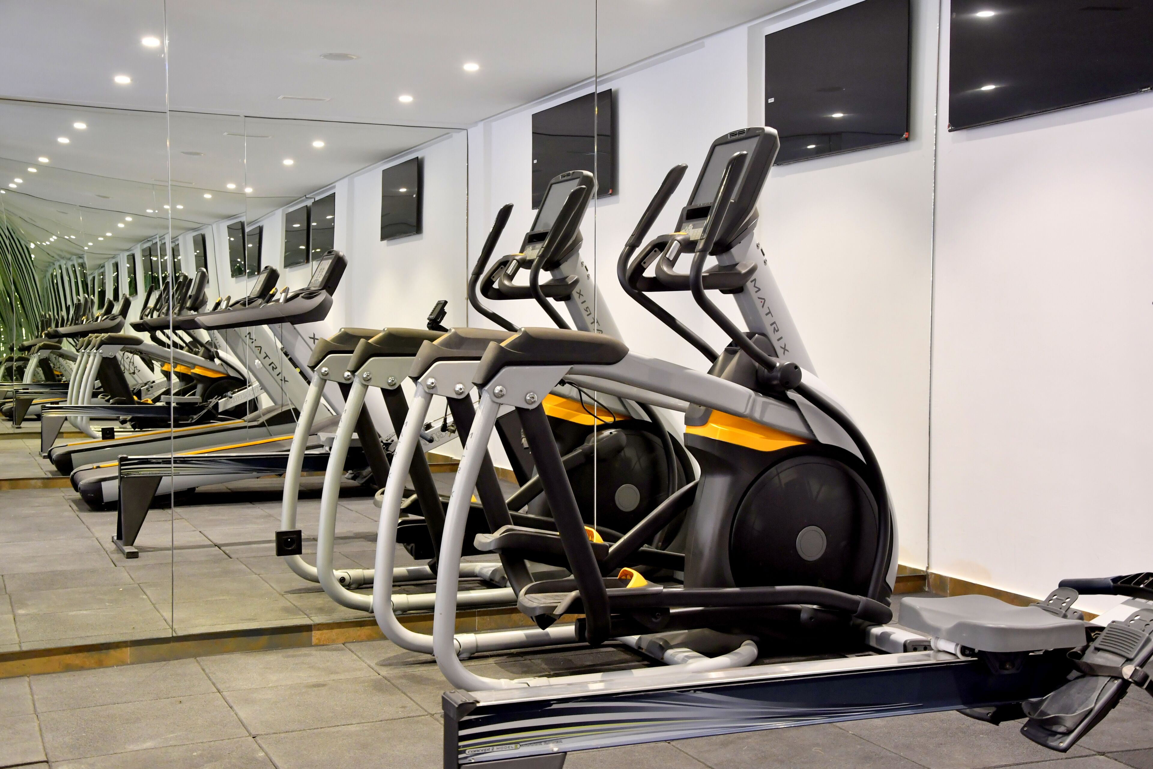 Une rangée de vélos d’appartement dans une salle de sport aux murs et au plafond recouverts de miroirs, éclairée par des luminaires.
