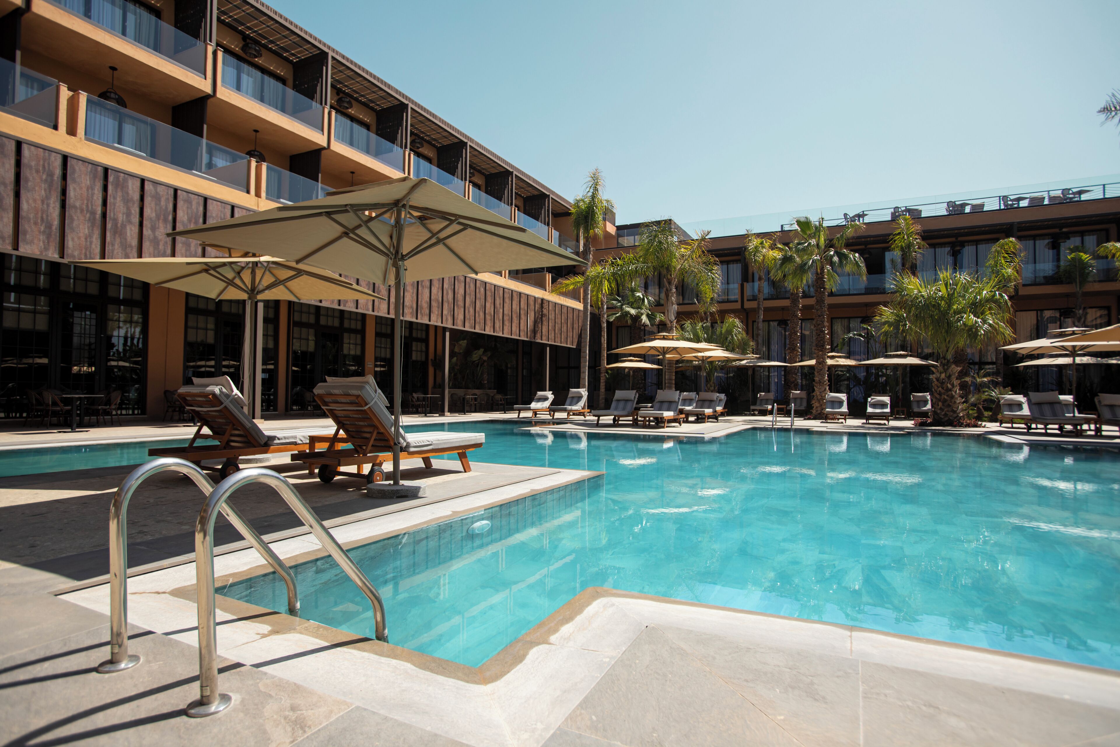Un área de piscina con sillones de descanso y sombrillas, un bar junto a la piscina y un edificio de varios pisos con balcones.