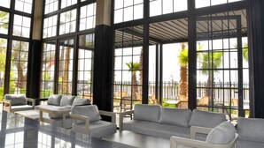 A spacious indoor area with large windows, multiple seating arrangements, and a view of palm trees outside. - Palm Taghazout Hotel (Tamraght)