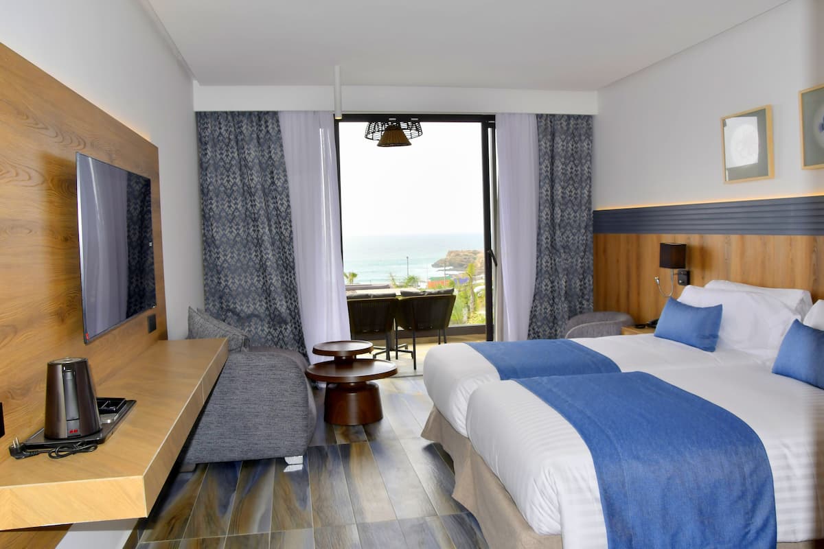 A hotel room with a bed, a TV, a coffee maker, a small table, and a view of the ocean.