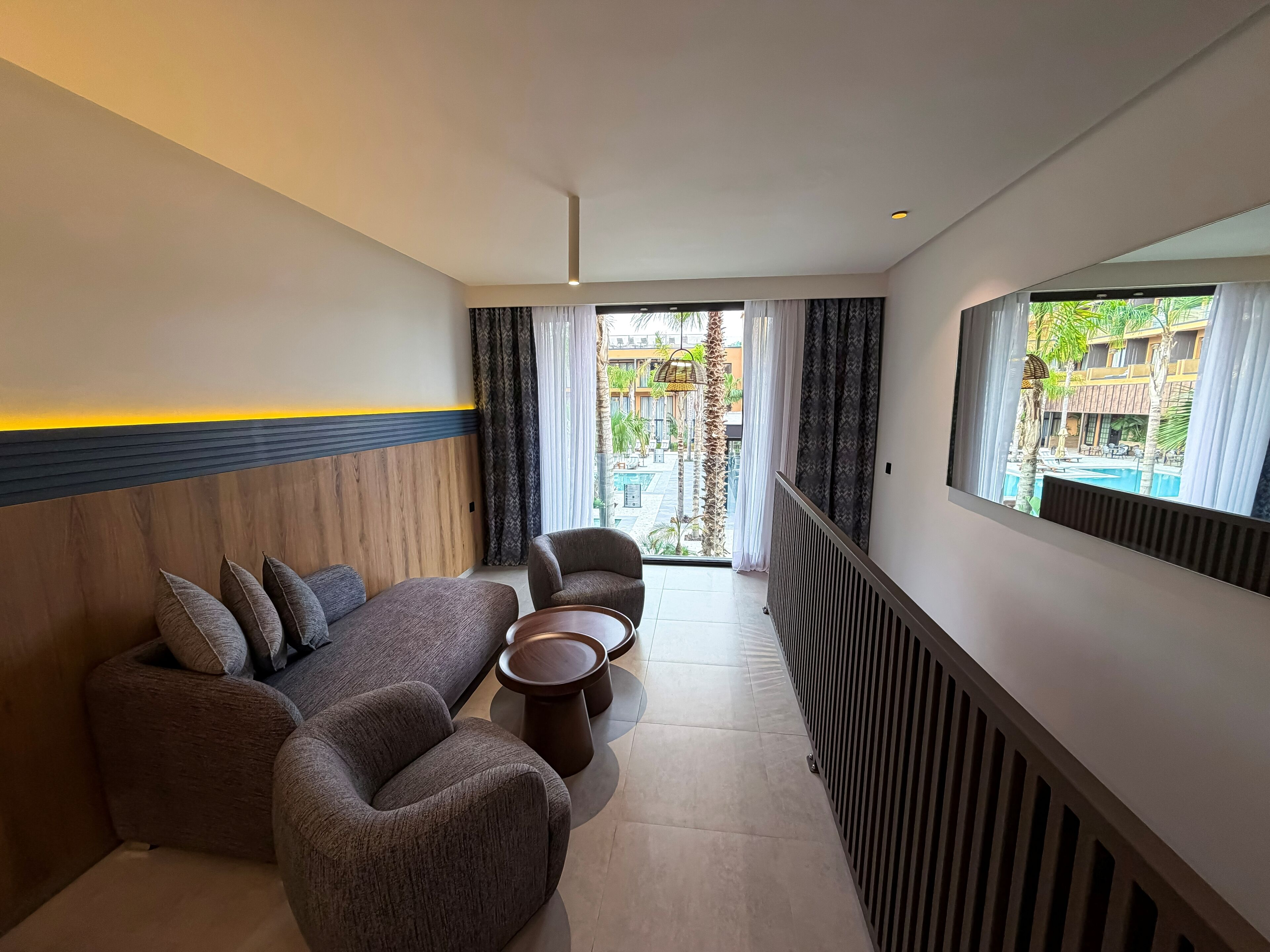 A modern hotel room with a sofa, armchair, and coffee table, a large window with a view of a pool and buildings, and a ceiling with recessed lighting.