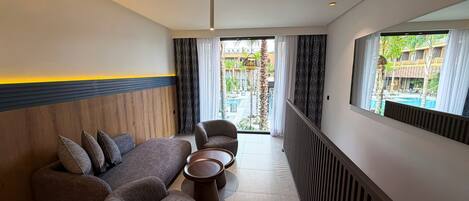 Una habitación de hotel moderna con sofá, sillón y mesa de centro, un ventanal con vistas a una piscina y edificios, e iluminación empotrada en el techo.