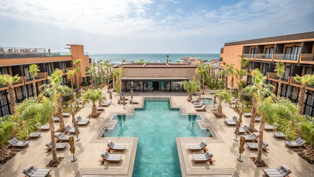 A hotel pool area with multiple lounge chairs and palm trees, surrounded by buildings with balconies overlooking the ocean. - Palm Taghazout Hotel (Tamraght)