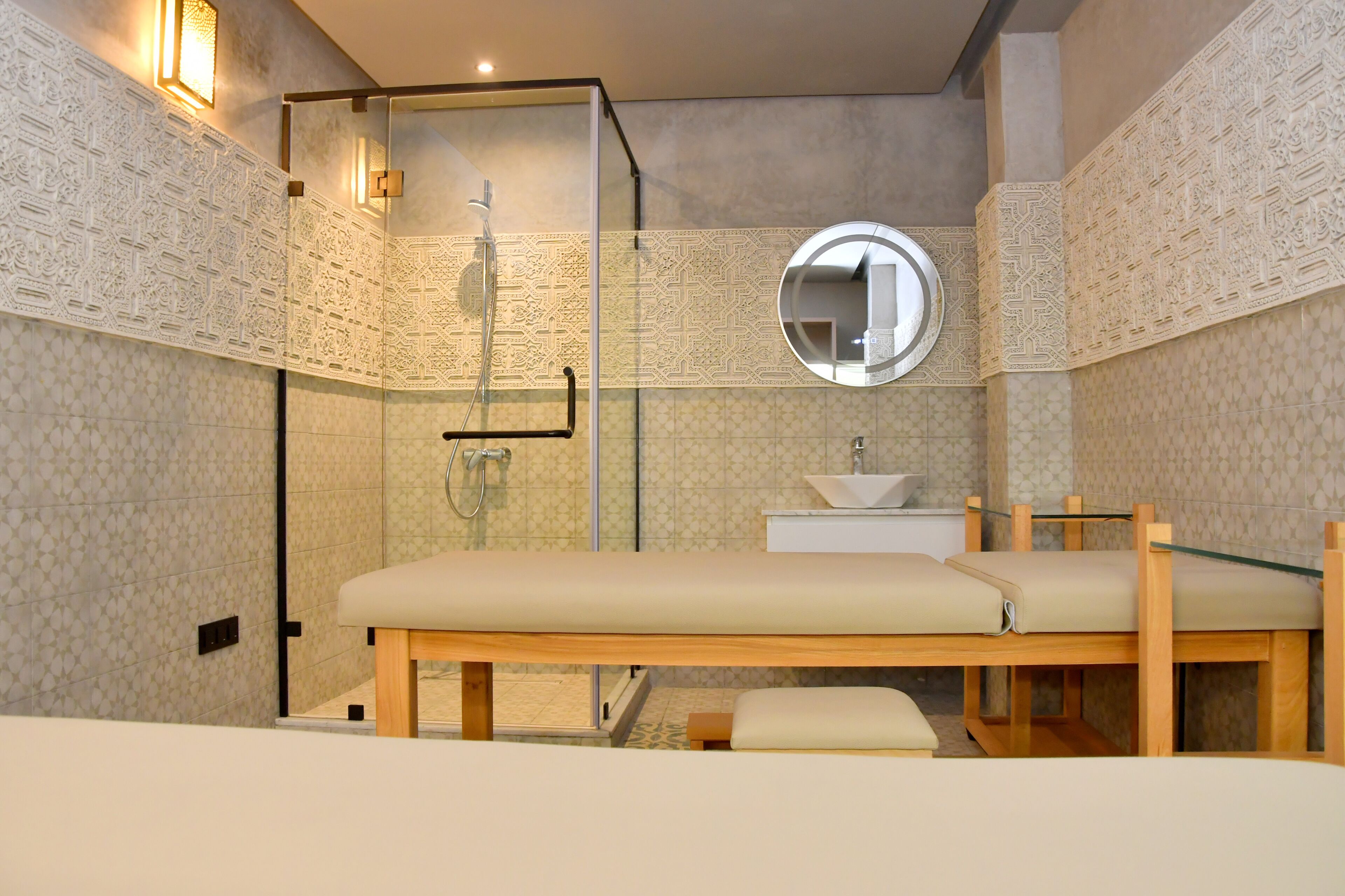 A modern bathroom with a glass shower enclosure, a round mirror, a sink, and a bench.