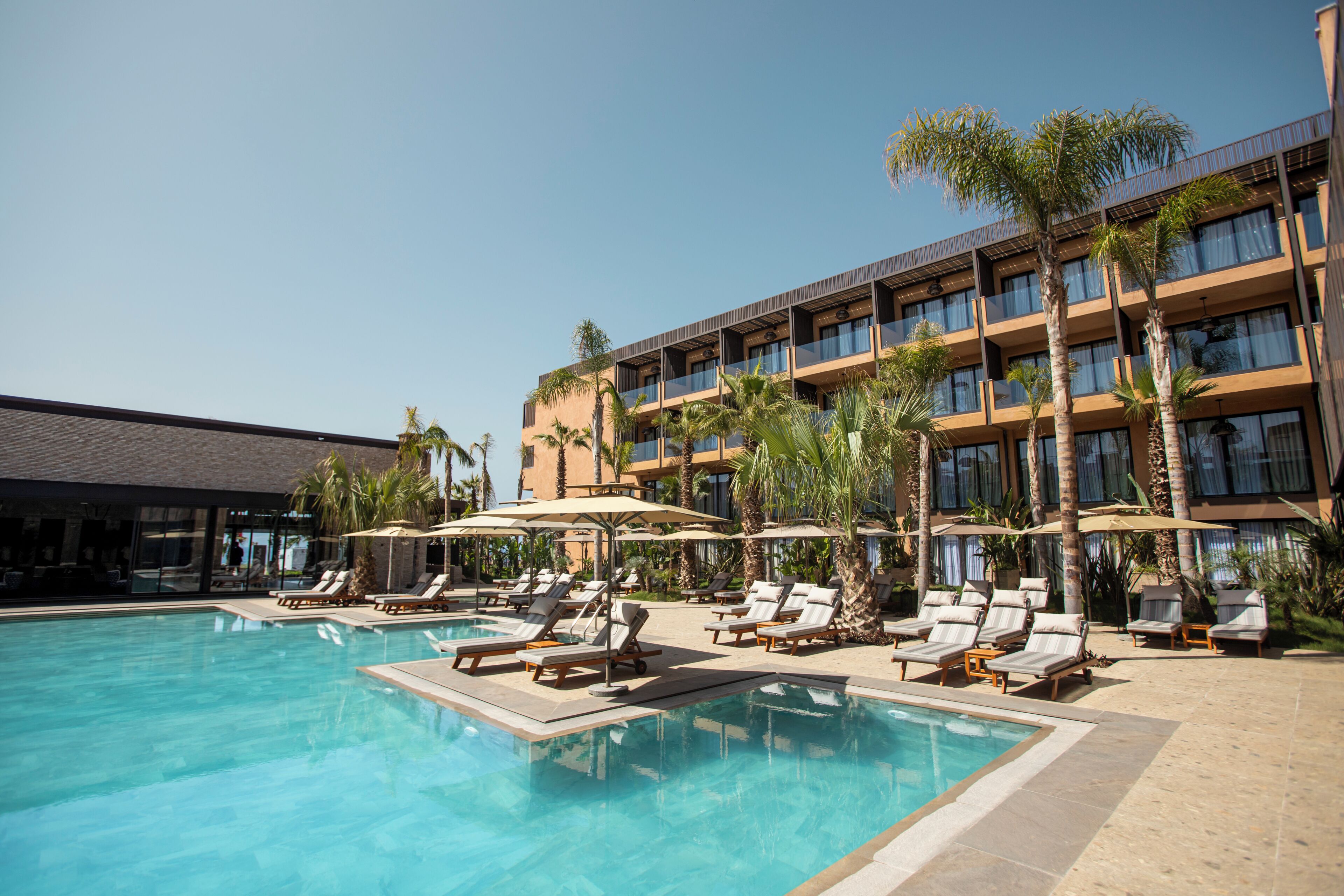 A pool area with lounge chairs and umbrellas, a building with multiple balconies, and palm trees.