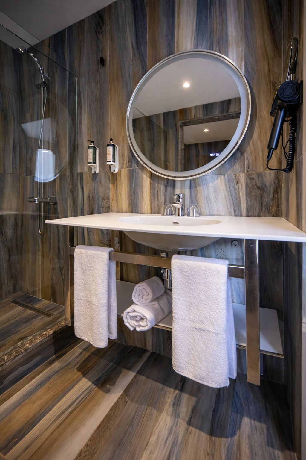 A modern bathroom with a wooden wall, a large mirror, a sink, and a towel rack.