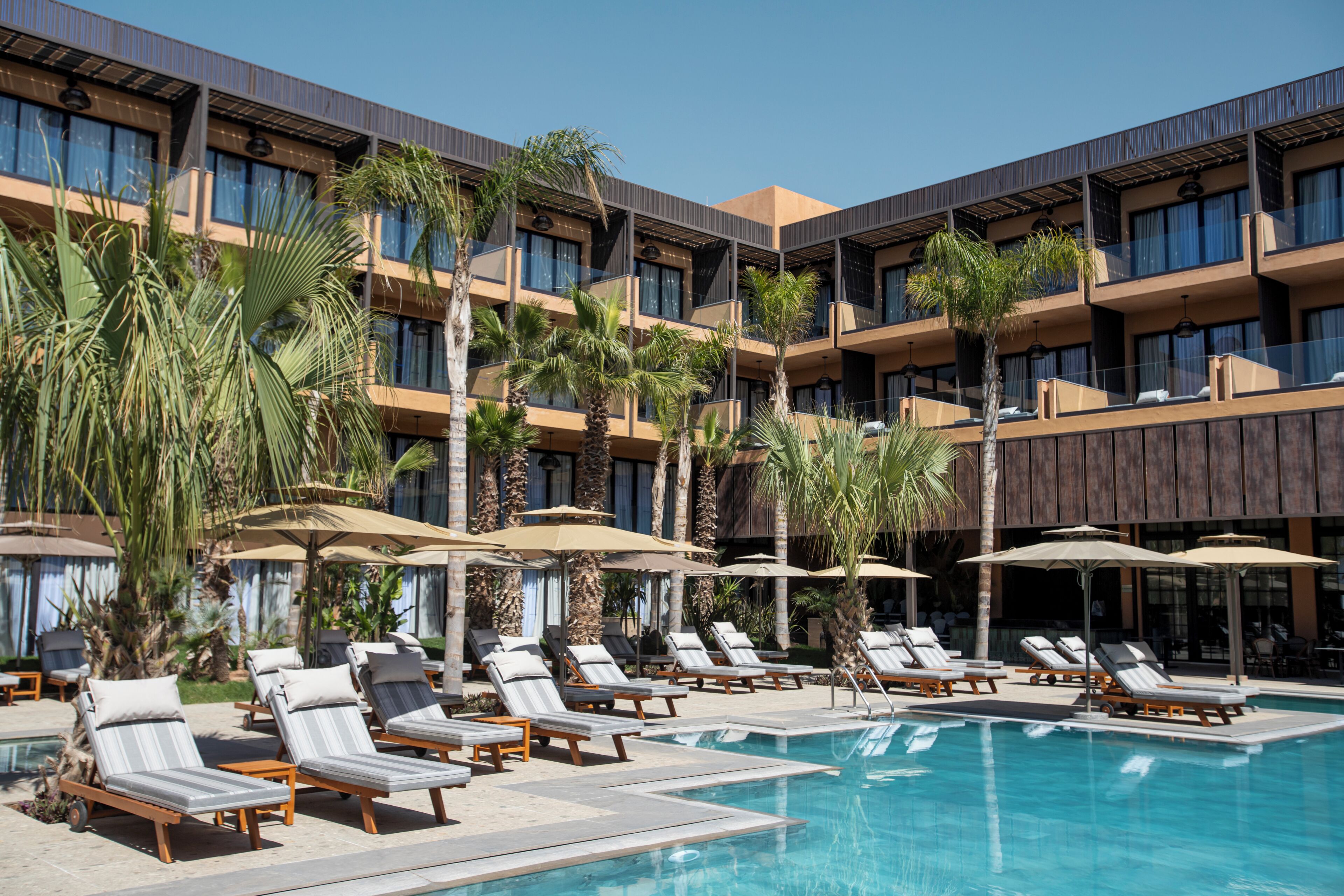 A hotel with a pool area, sun loungers, umbrellas, and palm trees.