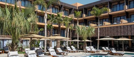 A hotel with a pool area, sun loungers, umbrellas, and palm trees.