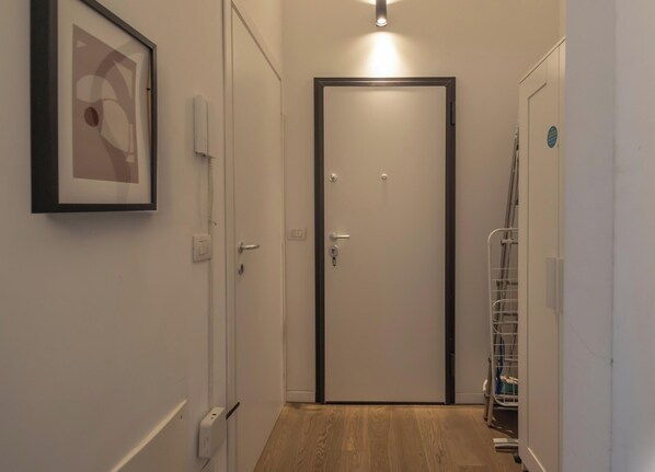 En hall med ett inramat konstverk på vänster sida, en dörr med en svart ram längst ner och ett metallställ med diverse föremål på höger sida.