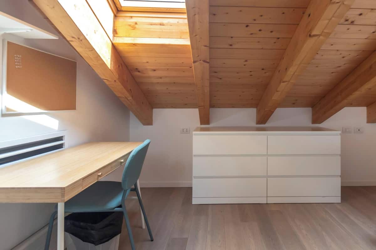 A room with a sloped ceiling, a desk, a chair, a white dresser, and a skylight.