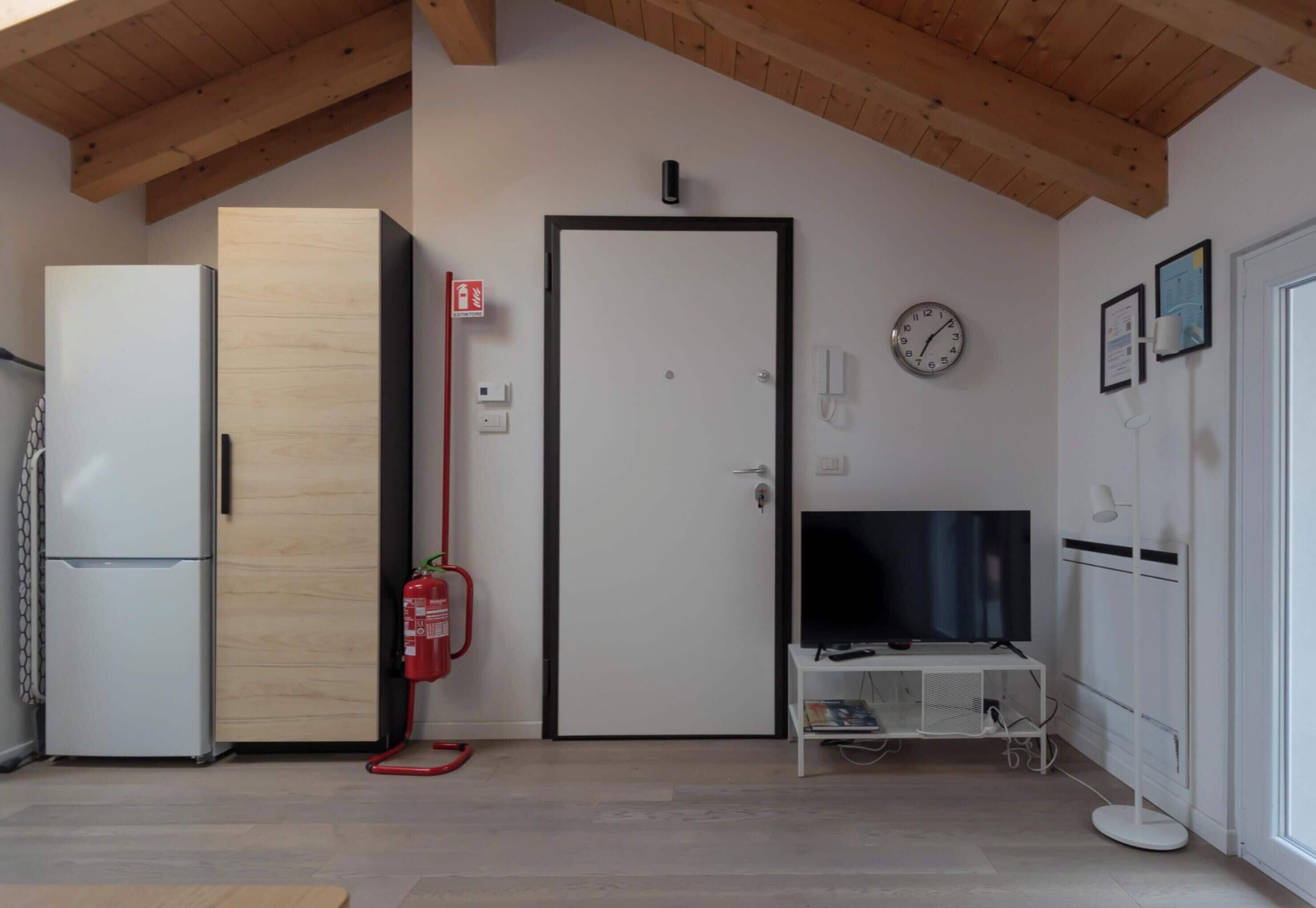 Una habitación con una puerta blanca, un mueble blanco para televisor, un refrigerador blanco, un armario de madera, un extintor y un reloj en la pared.