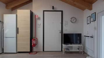 Una habitación con una puerta blanca, un mueble blanco para televisor, un refrigerador blanco, un armario de madera, un extintor y un reloj en la pared.