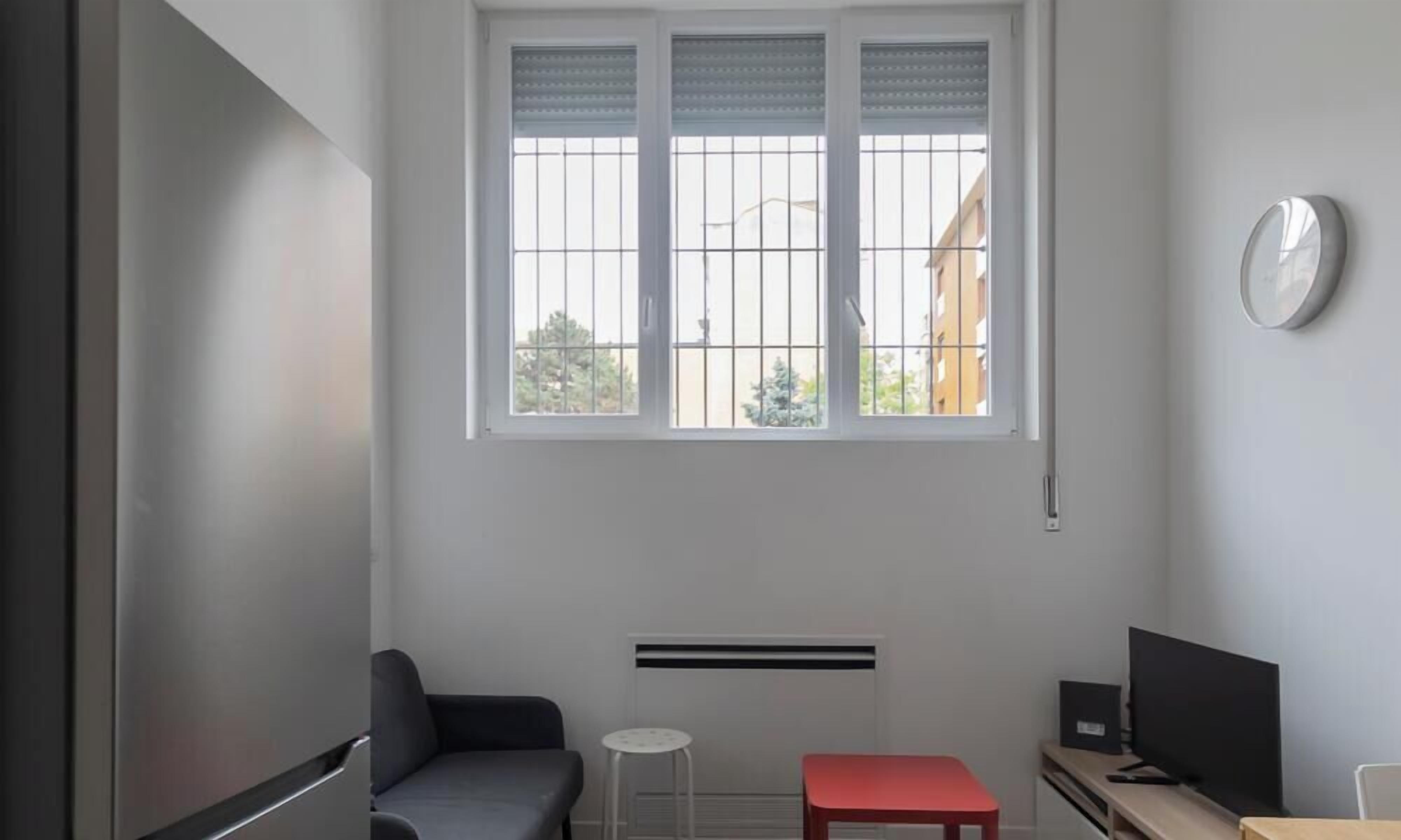 A room with a window, a radiator, a small table, a red bench, and a TV.