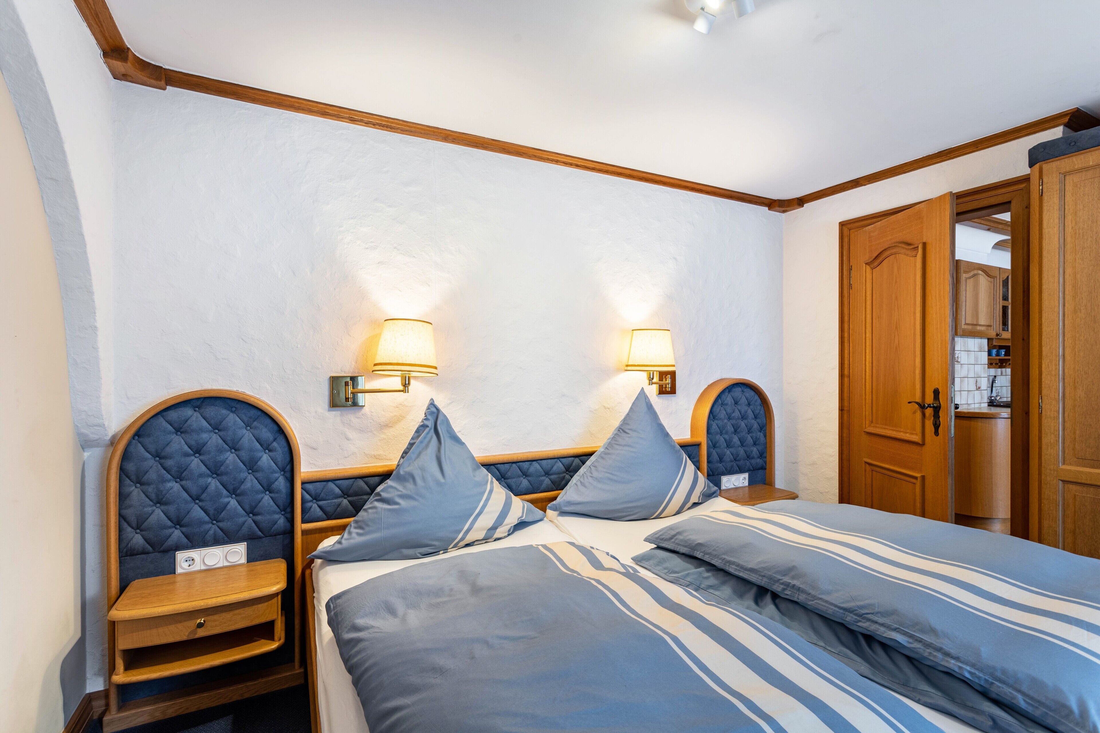 A bedroom with two beds, nightstands, wall-mounted lamps, and a wooden wardrobe.