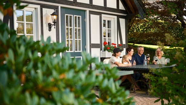 Eine Familie speist draußen an einem Tisch mit Blick auf ein traditionelles Haus mit freiliegenden Holzbalken und einer dunkelblauen Tür.