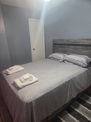 A bedroom with a bed, pillows, folded towels, a door, and a carpeted floor.