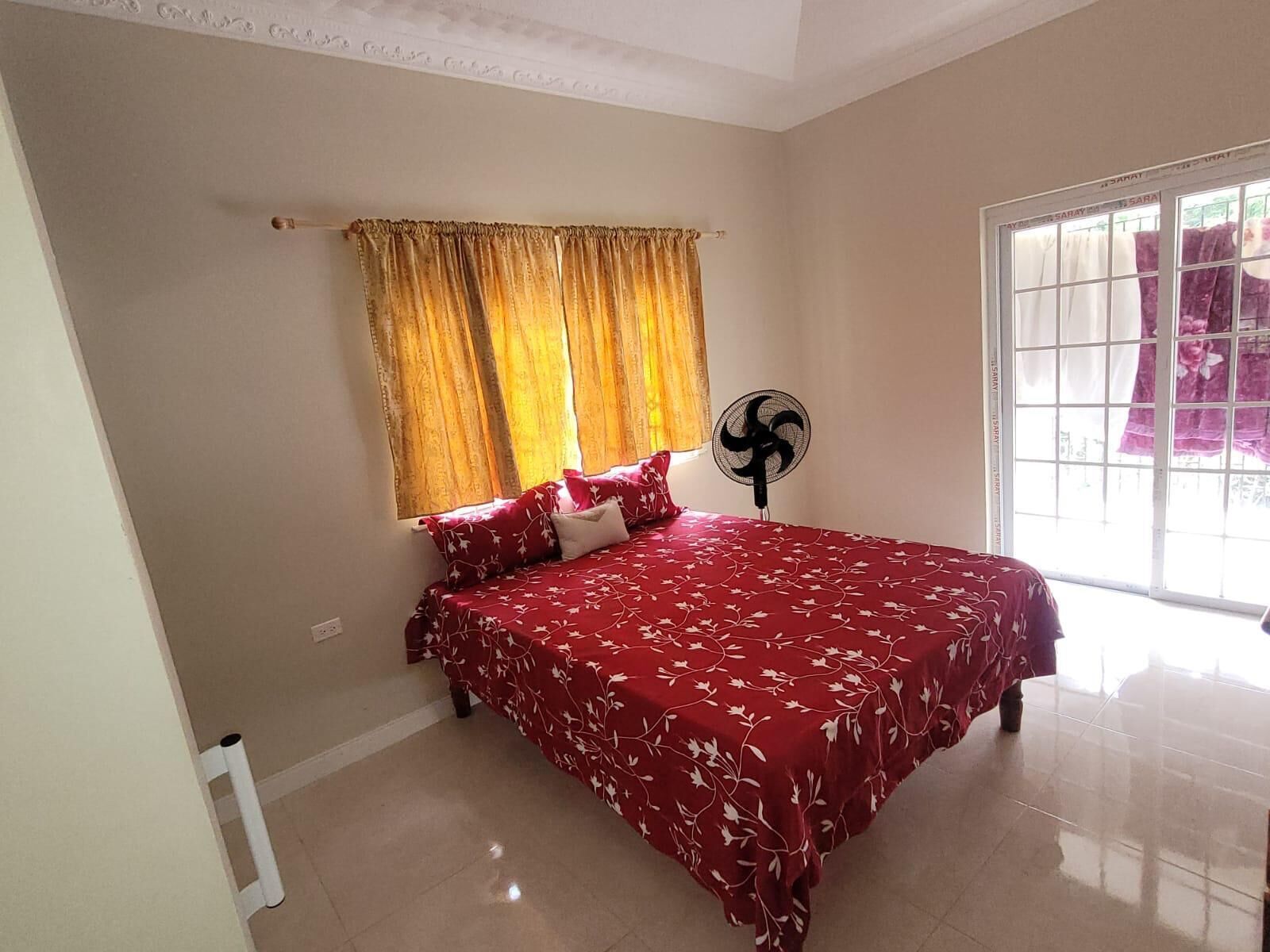 A bedroom with a bed, a window with curtains, a standing fan, and a radiator.