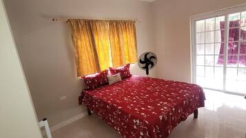 A bedroom with a bed, a window with curtains, a standing fan, and a radiator.