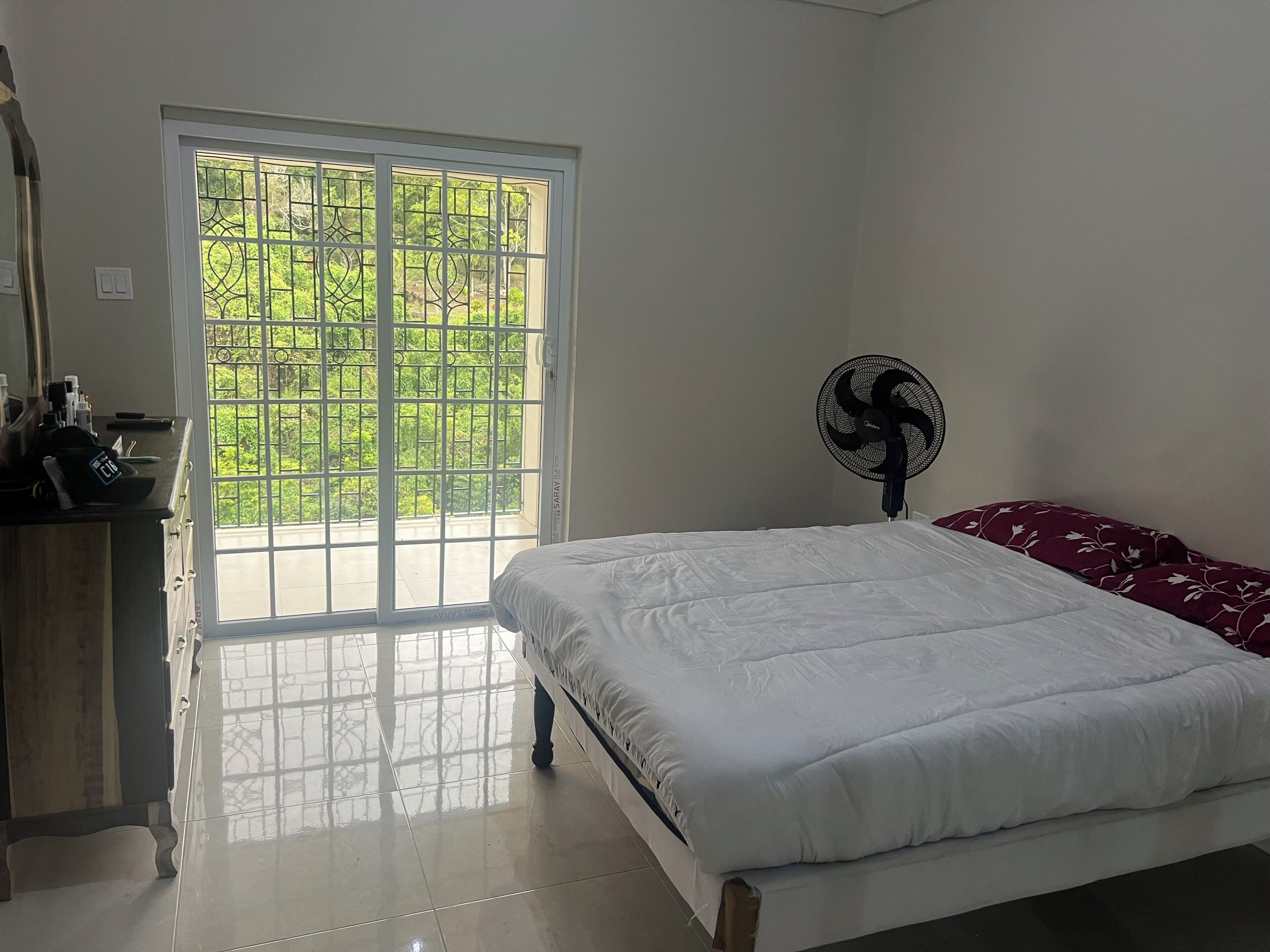 A bedroom with a bed, a fan, a dresser, and a window with a view of greenery.