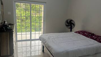 A bedroom with a bed, a fan, a dresser, and a window with a view of greenery.