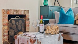A cozy living room with a brick fireplace, a vintage suitcase, a bottle of wine, and a stack of firewood. - Maple Cottage in Henley (Henley-On-Thames)