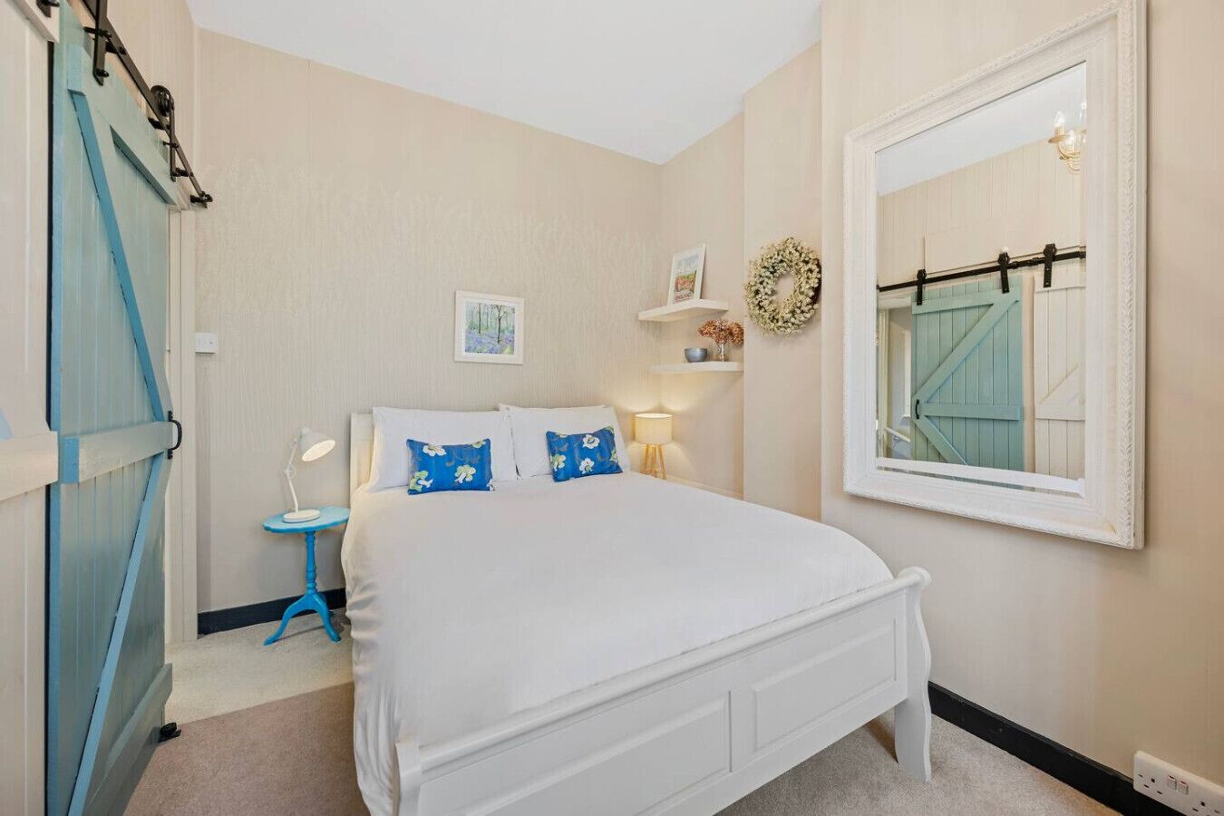 Ein Schlafzimmer mit einem weißen Bett, einem kleinen Nachttisch, einem wandmontierten Spiegel und einer blauen Tür.