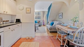 A cozy kitchen with white cabinets, a dining area with a rustic wooden table, and a living room with a sofa and blue cushions. - Maple Cottage in Henley (Henley-On-Thames)