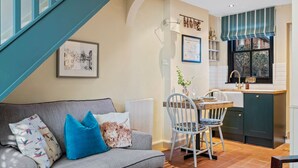 A cozy living room with a grey sofa, blue cushions, and a dining area with a wooden table and chairs. A kitchen with dark cabinets and a window with a striped valance is visible. - Maple Cottage in Henley (Henley-On-Thames)