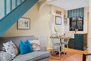 Una sala de estar acogedora con un sofá gris, cojines azules y un comedor con una mesa y sillas de madera. Se ve una cocina con armarios oscuros y una ventana con un cortinero a rayas.