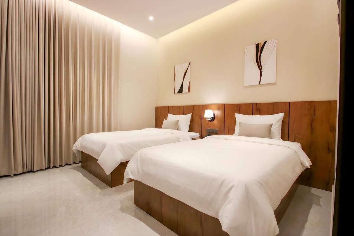 A hotel room with two beds, a wooden headboard, a large window with curtains, and two wall-mounted light fixtures.