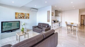 A modern living room with a flat-screen TV, leather sofas, a dining area with a table and chairs, and a kitchen with cabinets and a stove. - Mellieħa 2BR Apt | Rooftop Hot Tub & View (Mellieħa)