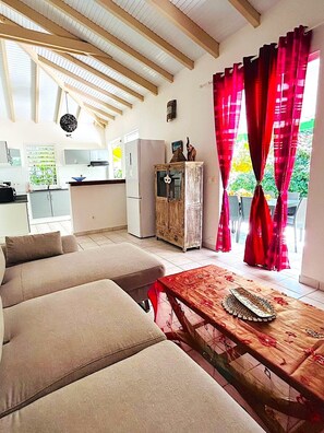 A cozy living room with a sofa and a wooden coffee table, a kitchen with white cabinets and a refrigerator, and a dining area with red curtains. - Villa Annia (Saint-François)